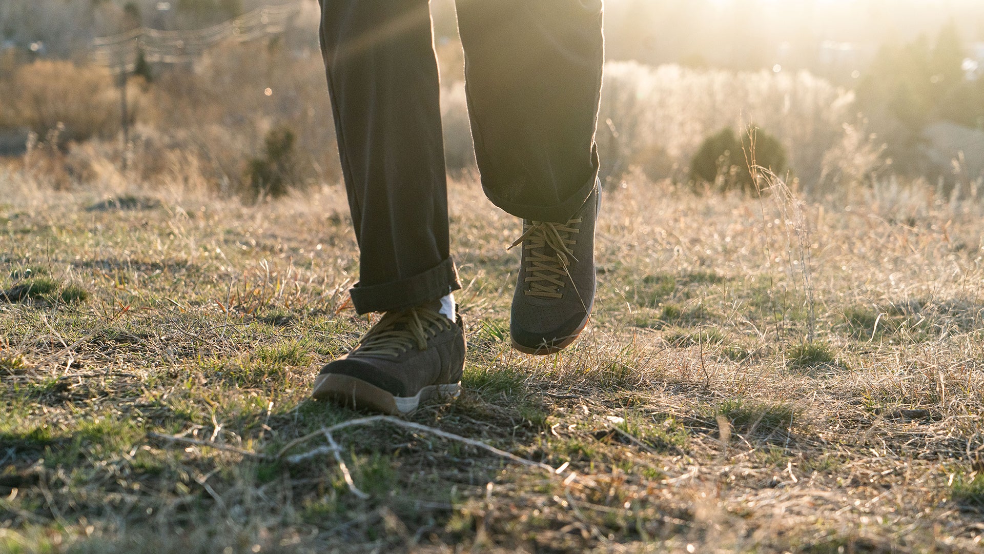 A person walking outdoors on dry grass during golden hour, wearing  brown Bozeman II  shoes and dark pants with a bright sun flare in the background.
