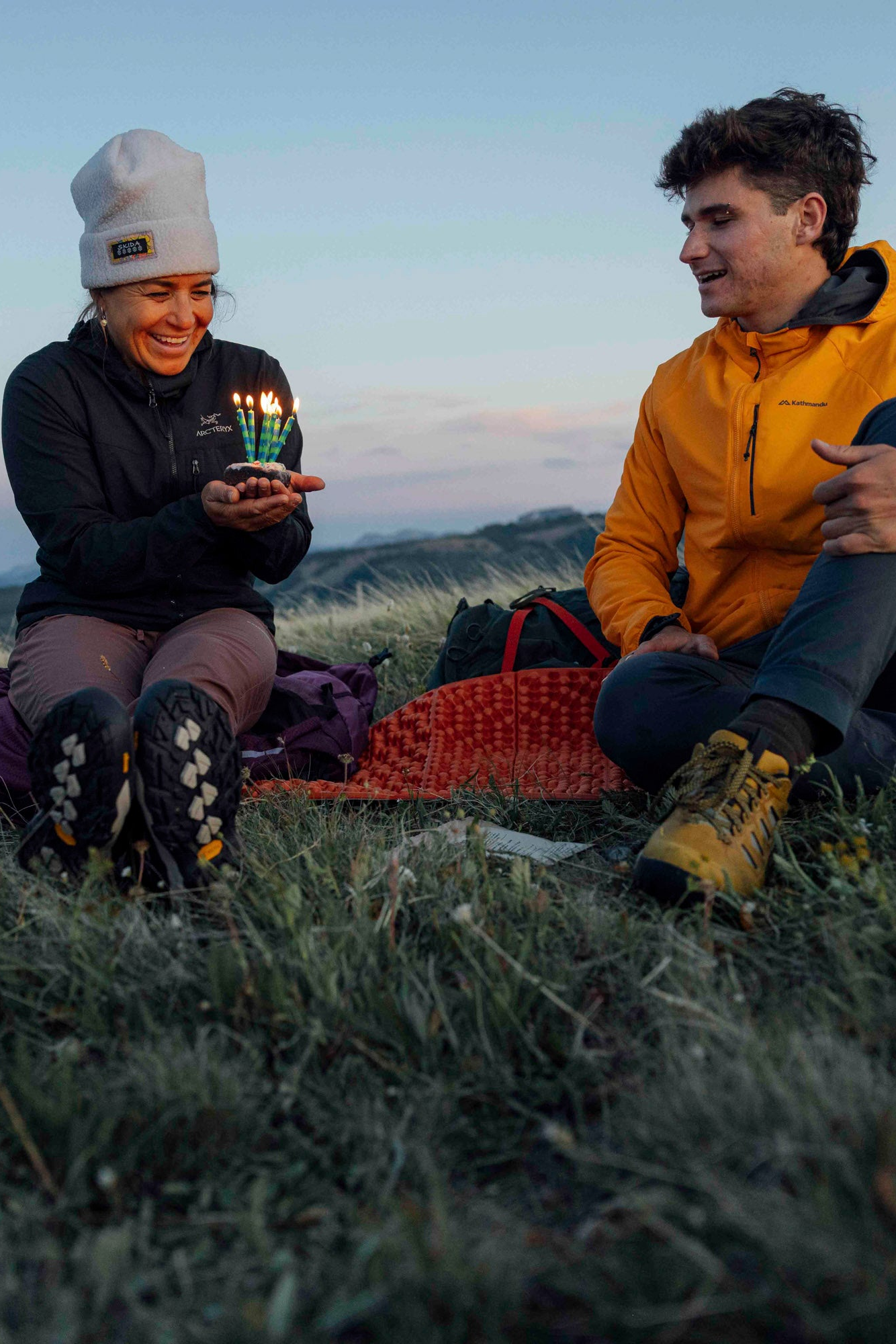 Two people sitting near a tent in a field at dusk.