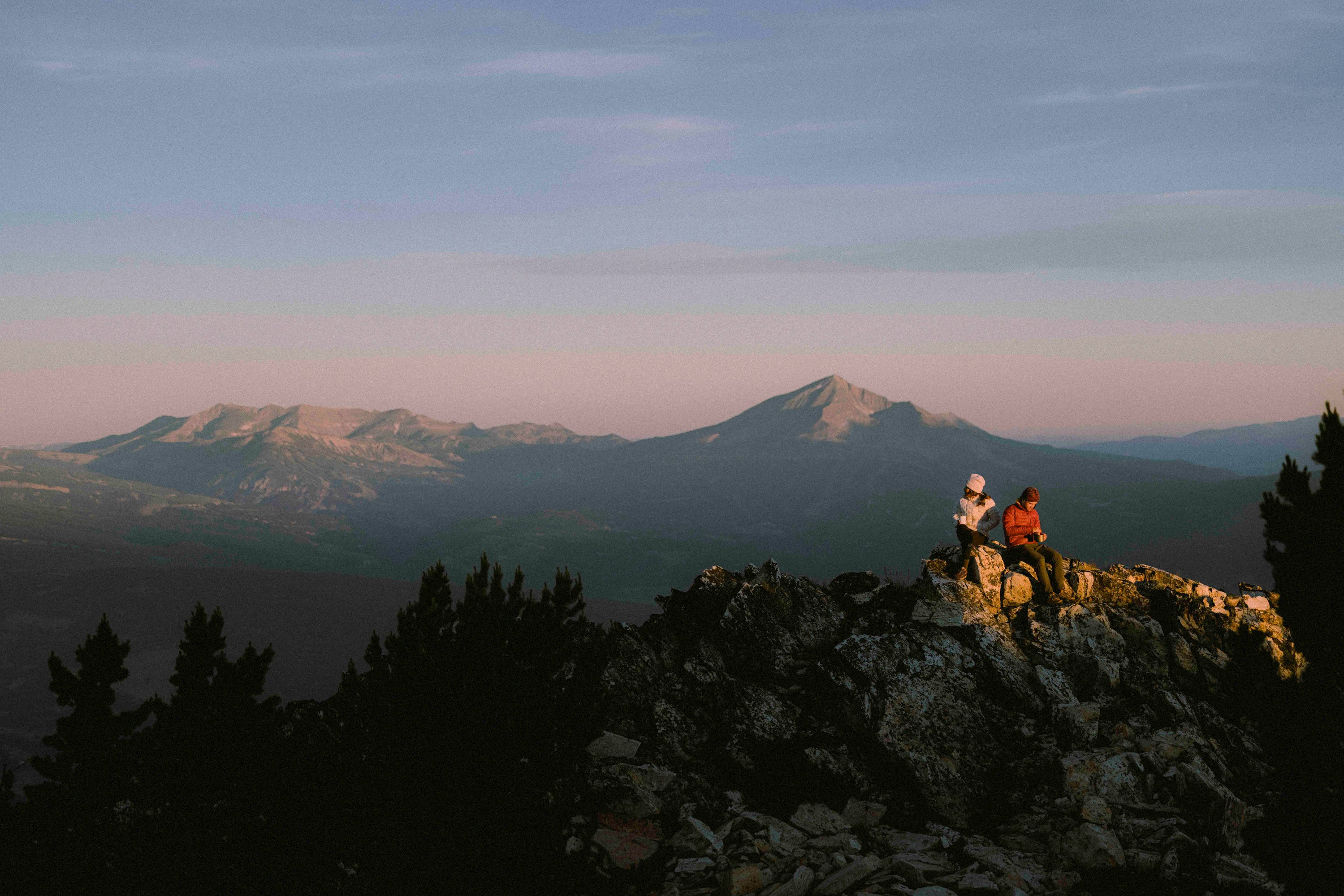 Two people sitting on a mountain top with a scenic view of mountains in the background.