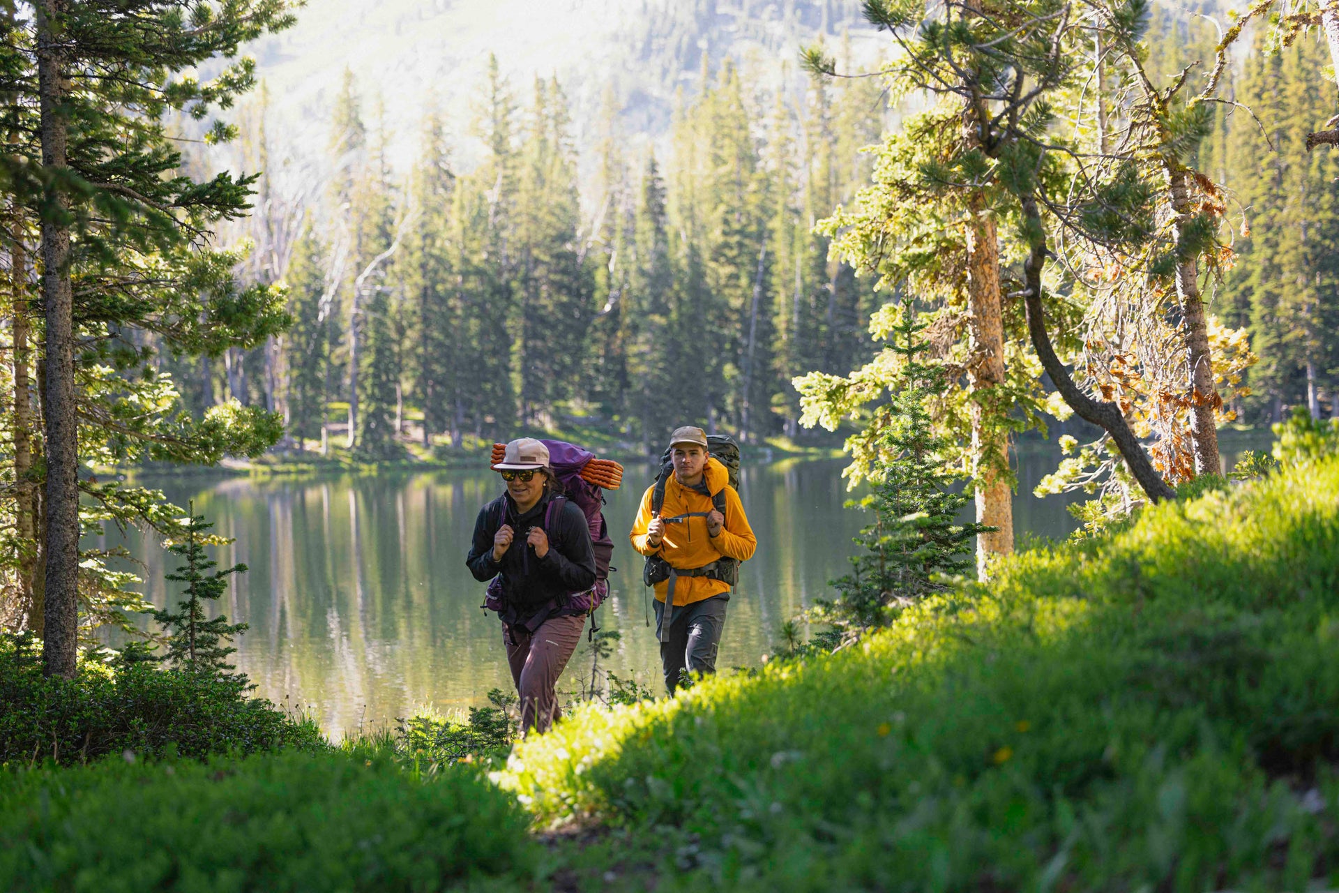 Two hikers with backpacks walking along a trail by a lake surrounded by trees.