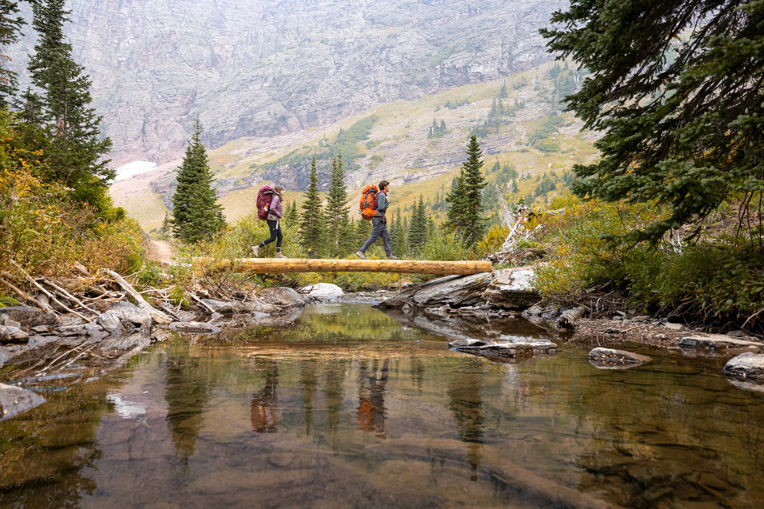 Two hikers with backpacks crossing a wooden bridge over a stream in a mountainous area.