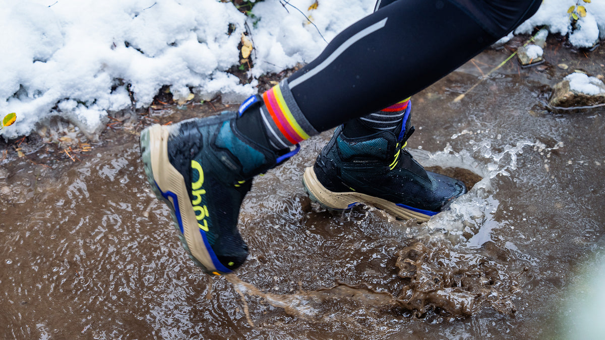 A close-up of a person's legs stepping into a muddy puddle outdoors, wearing black Oboz Katabatic Storm winter boots with yellow and blue accents, rainbow-striped socks, and black leggings with a white stripe.